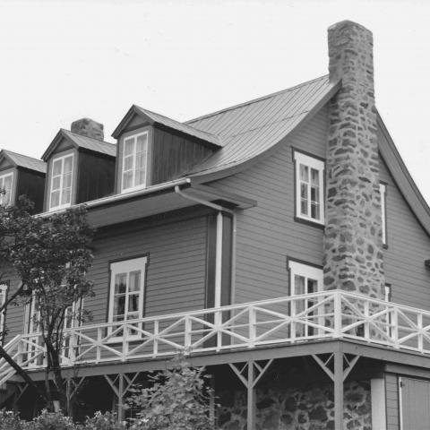 Solide maison québécoise ancienne, avec cheminée de pierre, toit en tôle et murs de planches horizontales en bois.