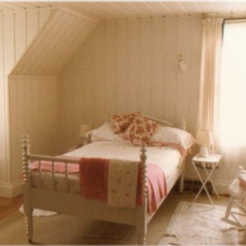 Chambre où se trouvent un lit à une place dont la base est en bois tourné, une commode, une berçante et un petit bureau.
