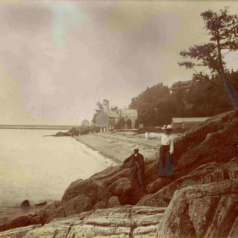 A couple of vacationers have climbed onto the rocks; a beach and a long wharf are in the background.