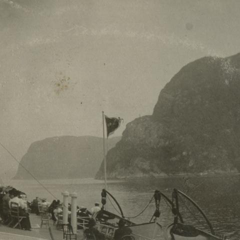Passengers sitting on the bridge of a steamer, looking at the steep mountains on the shore.