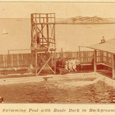 Many people sitting around a fenced pool with a large diving platform.
