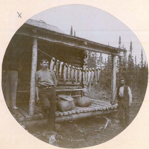 Two men pose beside a dozen fish suspended between posts supporting the porch roof at a fishing camp.