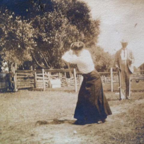 A woman swinging to hit a golf ball while a man waits in the background.