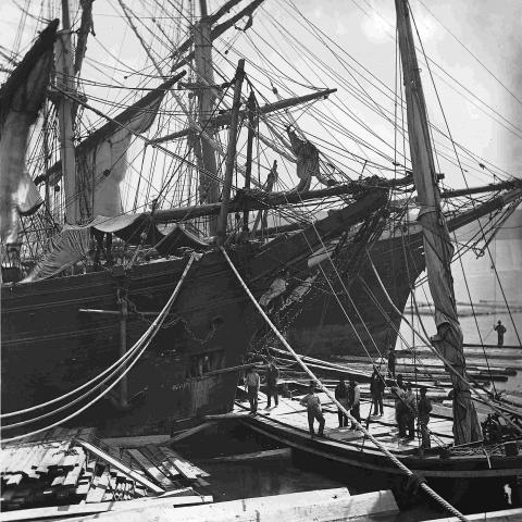 A dozen men unloading a large sailing vessel transporting lumber.