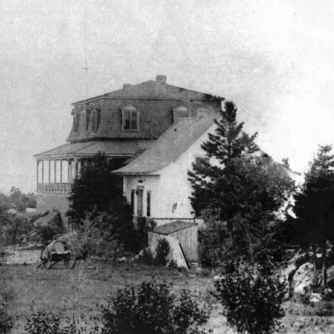 Photographie ancienne d’une maison de campagne entourée d’arbres, avec une falaise en arrière-plan.