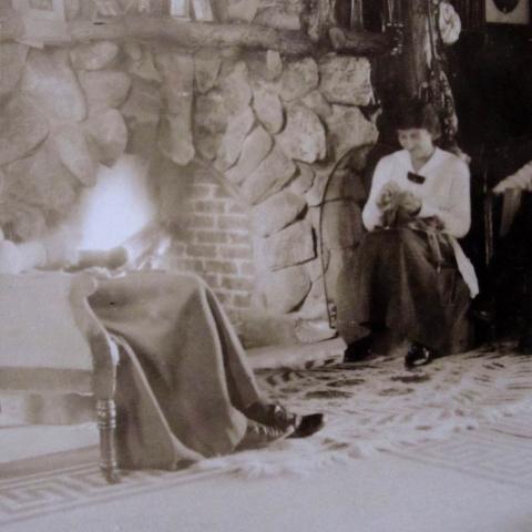 Three young women are reading and sewing in front of a fieldstone fireplace.