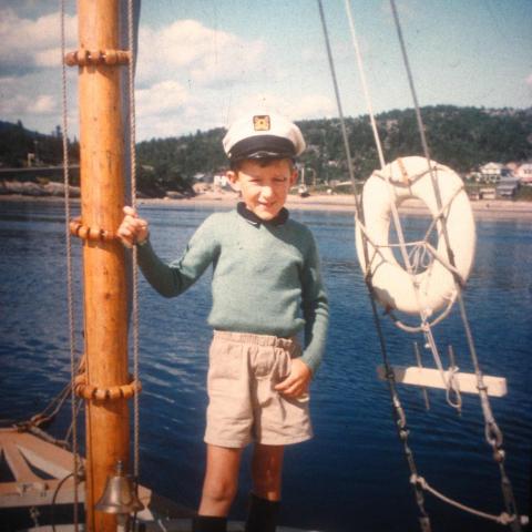 A colour photograph of a boy standing on the deck of a sailboat with the shore in the background.