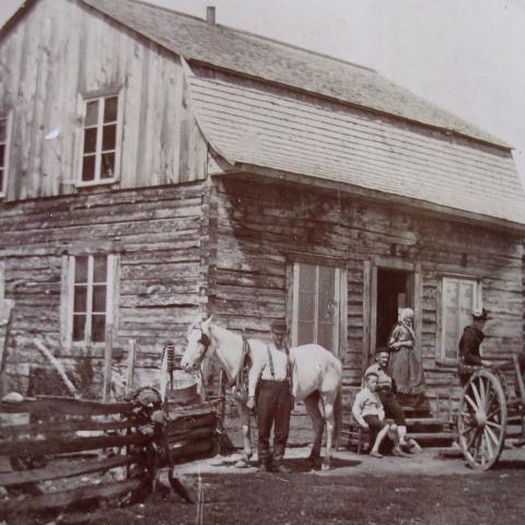 Quelques personnes posent devant une maison d'habitant construite en pièces sur pièces. Deux chevaux sont au repos.