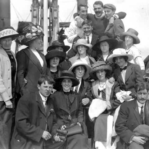 Un groupe d’hommes et de femmes; celles-ci portent des chapeaux à la décoration chargée.