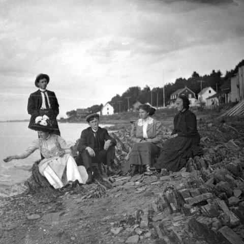 Cinq adultes, dont une dame âgée, posent sur une plage très rocheuse. Une femme semble lancer du pain à un oiseau hors champ.