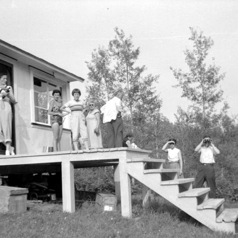 A dozen or so people behind a cottage in casual dress, two holding binoculars.