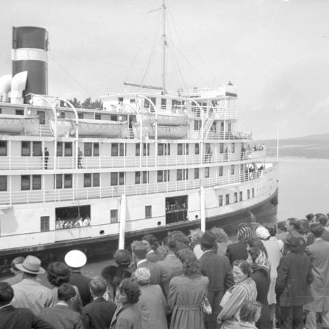 Dozens of passengers waiting on a wharf where a very large cruise steamer is moored.