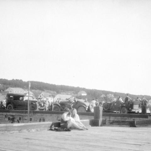 Two young women talking on an action-filled wharf, with several cars parked in the background.