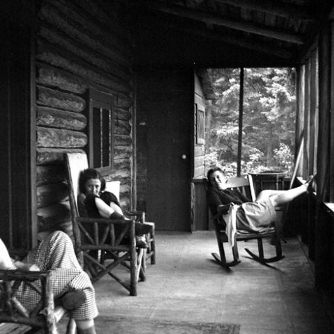 Three young women rocking on the porch of a log cabin.