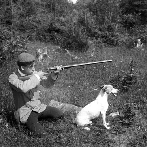 A young man is kneeling in the grass with his dog, hunting rifle in hand.