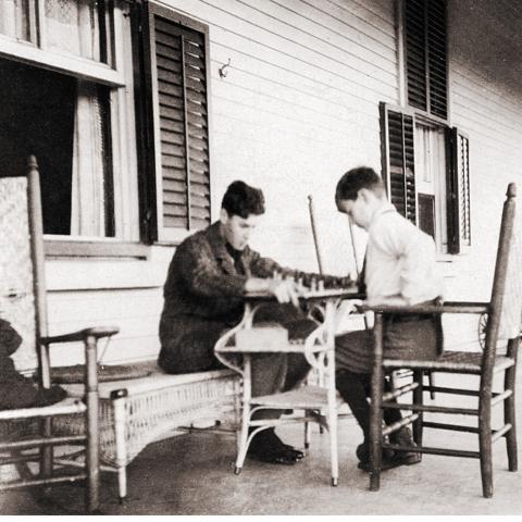 Two well-dressed teenage boys play chess on a veranda.