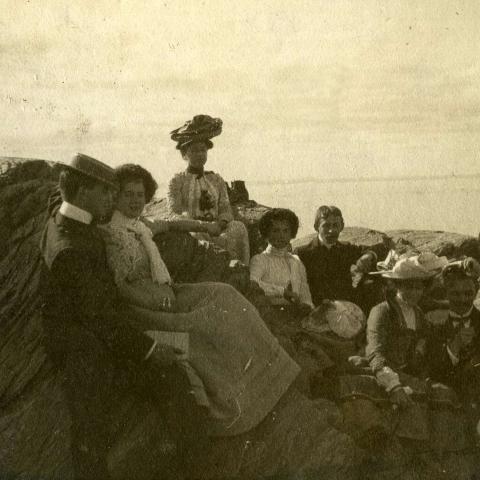 Trois couples de jeunes gens sont assis sur les rochers au bord de l’eau, accompagnés d’une femme et d’un jeune homme.