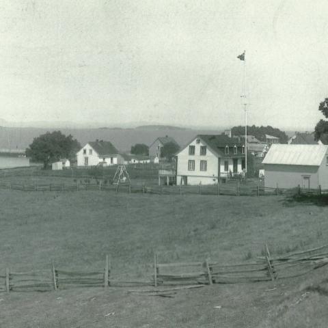 Beyond a field are a few houses and a wharf on the St. Lawrence River. The far shore is in the background.