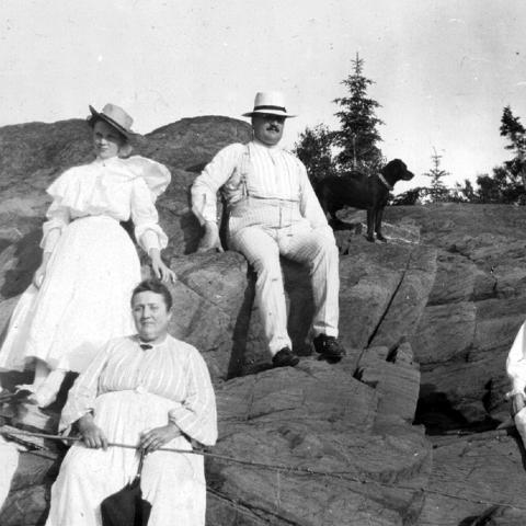 Five adults wearing white and sitting on a rock, with two dogs. One of the women is holding a fishing rod.