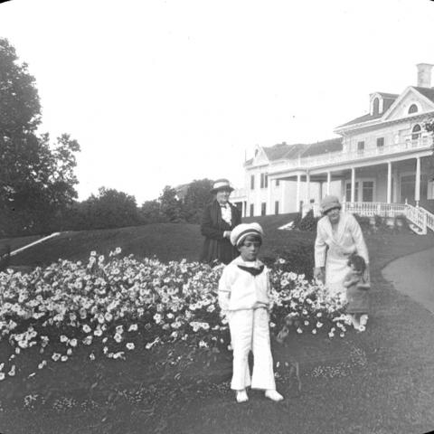 Two women and two children near flowerbeds in front of a very posh residence.
