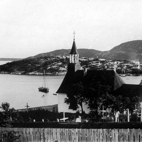 Une chapelle et son cimetière surplombent une baie, où sont amarrées des embarcations, dont deux bateaux à voile.