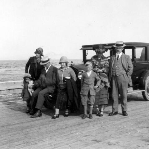 Cinq adultes et deux enfants en vacances posent devant une automobile ancienne stationnée sur un quai.
