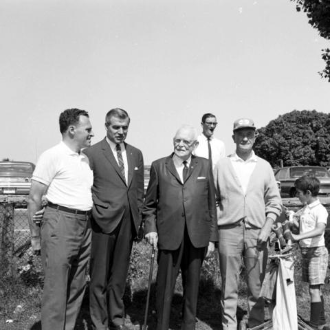 Portrait of a group of four men, followed by a caddie carrying a golf bag, with a parking lot in the background.