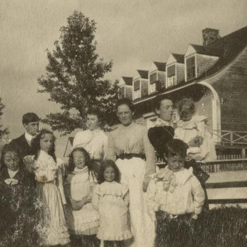 Three women and several children posing in front of a large old house with cedar shingling.