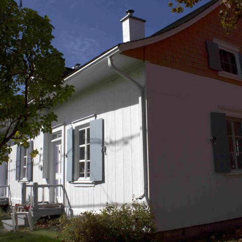 A colour photograph of an old white house with pale blue shutters, surrounded by trees.