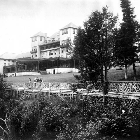 Un grand hôtel doté de grandes galeries. Des dames et de jeunes enfants observent le paysage près d’une balustrade.