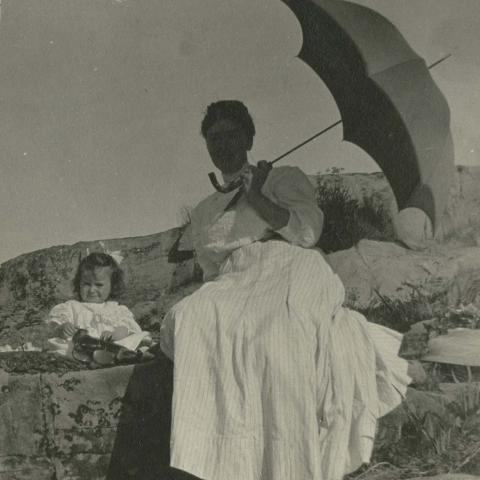 A women in a long dress and a young child sitting on a rock, with a parasol protecting them from the sun.