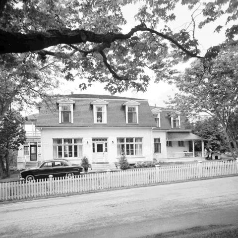 A white house with a mansard roof near a dirt road, with an automobile parked out front.