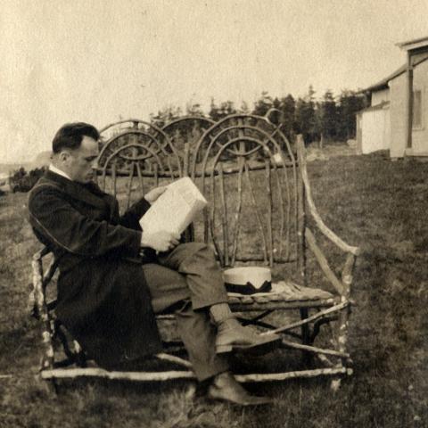 A man on vacation is reading, sitting on a wicker bench at the rear of a summer residence.
