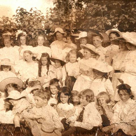 About thirty summer people of all ages, mainly young girls, posing by lilacs.