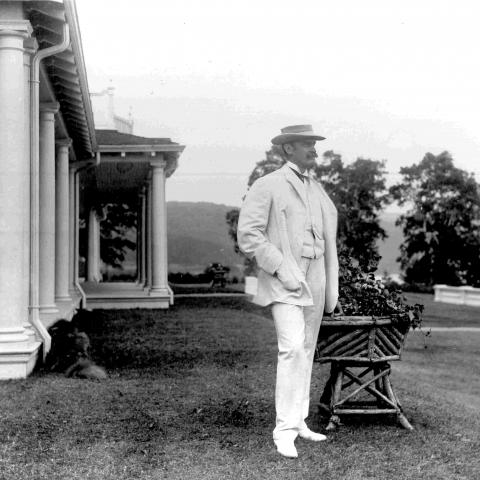 An elegant man wearing white and posing near an impressive residence, with a dog lying beside the house.