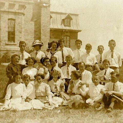 Some twenty elegantly-dressed young people pose in front of a castle-like building. A few of them hold tennis racquets.