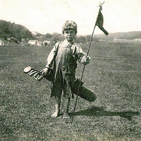 A barefoot child caddie, carrying a golf bag and holding a flag.