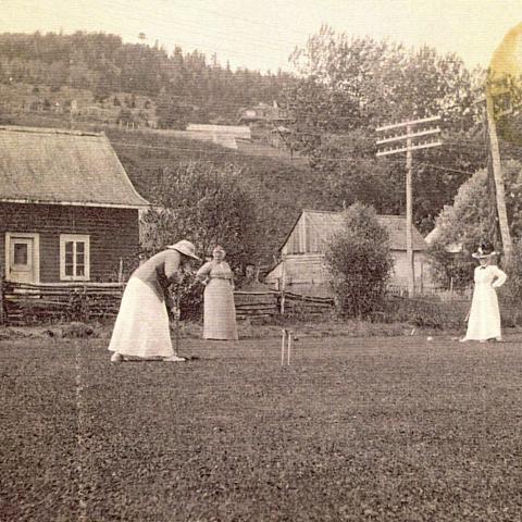 Three women play croquet near houses at the foot of a mountain.