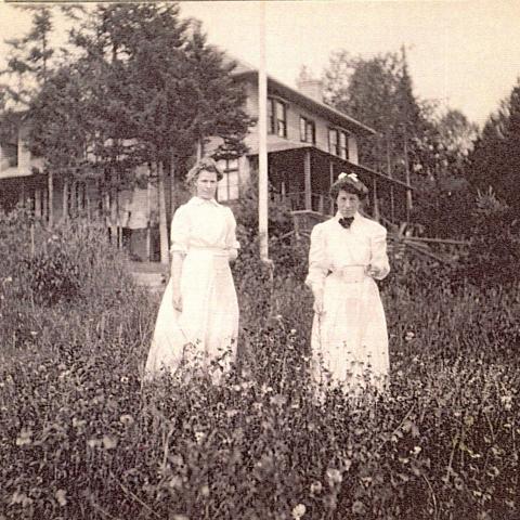 Two young women in dresses posing in a field near a house, each holding a white container.