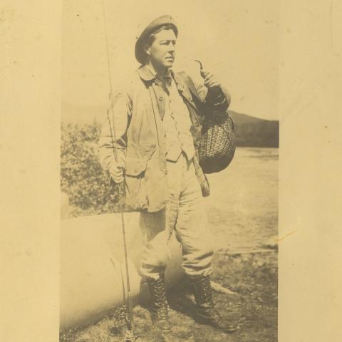 Photographie sur papier jauni d'un pêcheur et son équipement, dont une canne à pêche, un canot et panier à poissons en osier.