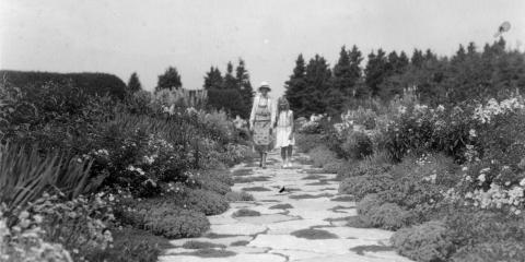 Une femme en tablier et une jeune fille marchent dans une l’allée fleurie d’un grand jardin.