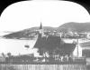 A chapel and its cemetery dominate a hill. Some houses are on the coast. A sailboat is moored.